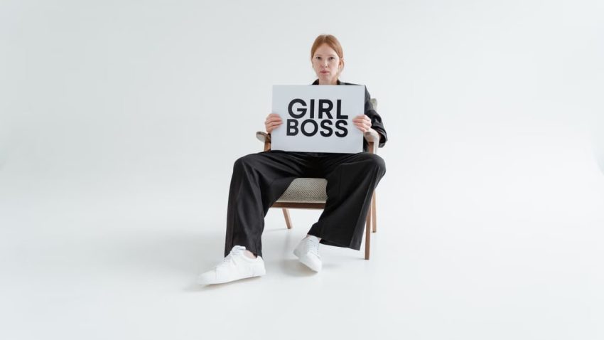 A woman in a black suit holds a 'Girl Boss' sign, exuding confidence on a plain background.